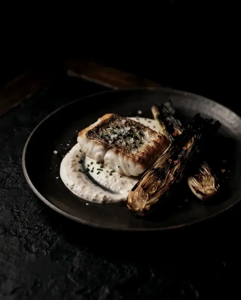 A roasted turbot fillet with crisp skin beside blackened charred leeks and mussel cream on a dark plate