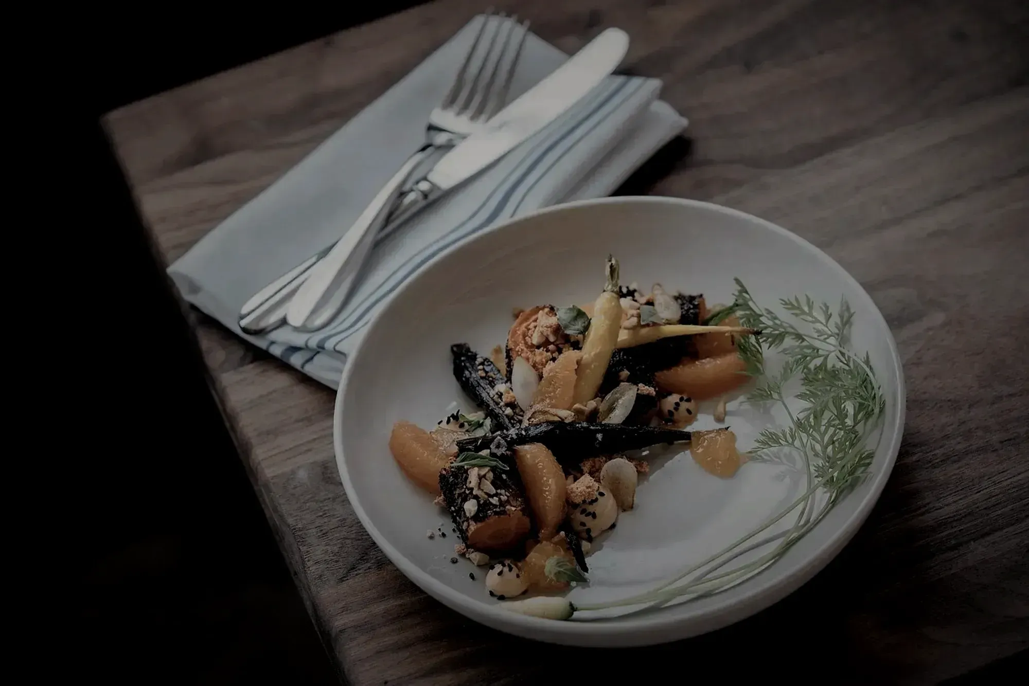 A plated autumn course of charred heritage carrots, blood orange and cultured cream on a pale bowl, lit low against a dark walnut table